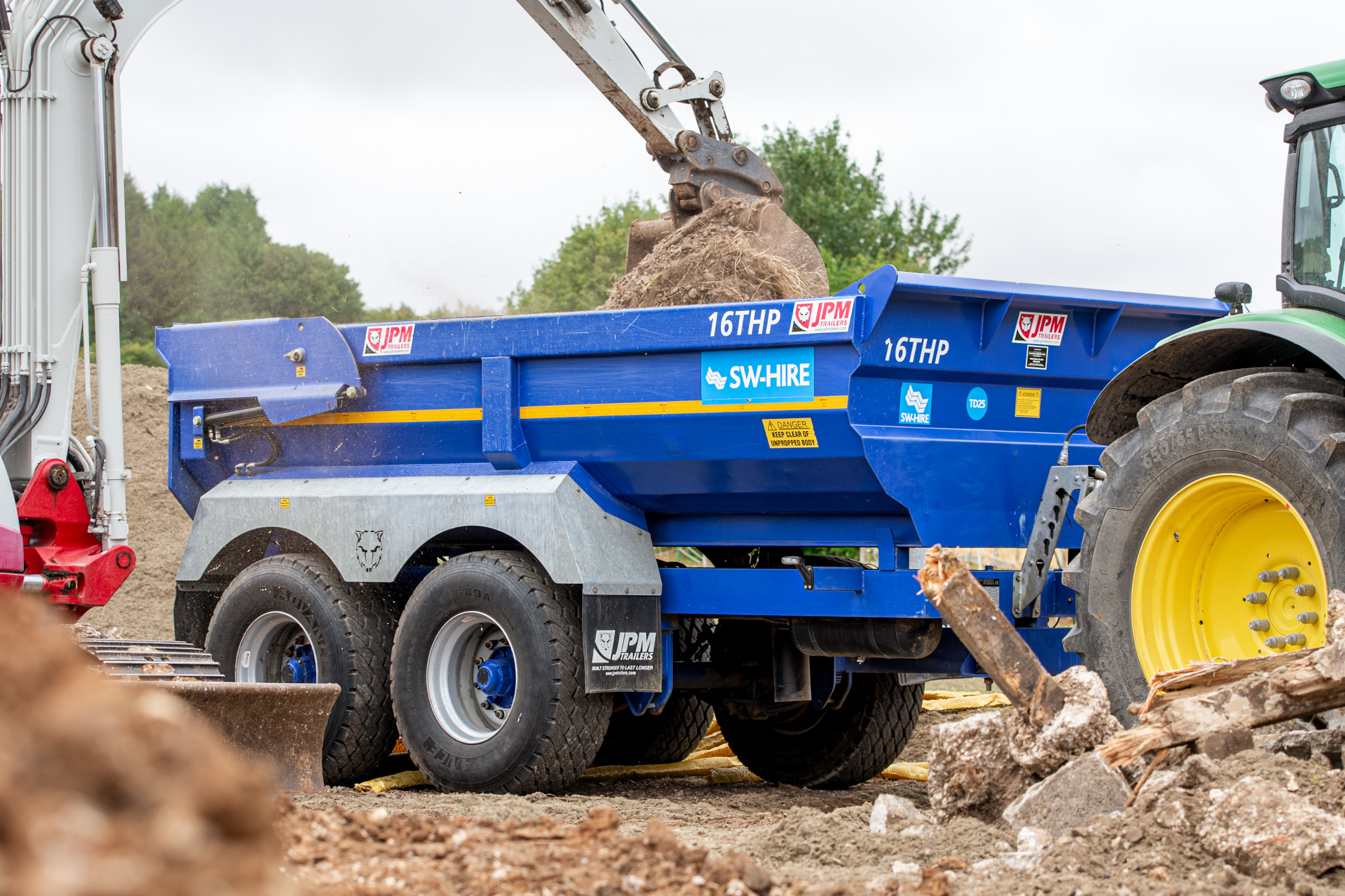 blue SW-Hire 16t JPM half-pipe dump trailer being loaded by a takeuchi excavator, towed by a john deere tractor